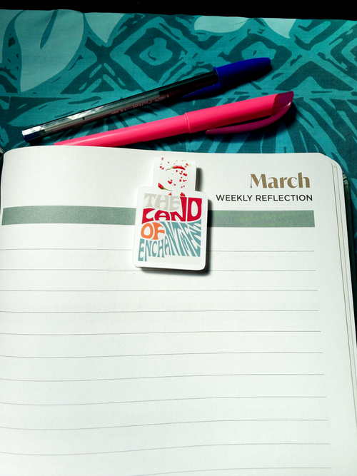 Magnetic bookmark with the words 'The Land of Enchantment' in a stylized, retro turquoise and red font. The design features small red chile pepper accents on a white background, clipped to a planner page.
